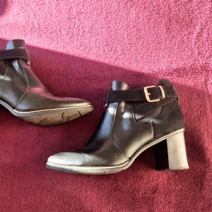 Svarta stövletter med klack och spänne från Star Collection- Stl 41  - Snygga svarta stövletter med rund tå och stabil klack. Stl 41. Skorna har en cool kombination av skinn och mocka, samt ett dekorativt spänne i metall på sidan. Dragkedja på insidan för enkel på- och avtagning. Perfekt för dig som gillar stilrena boots med lite edge. Använda en handfull gånger. 