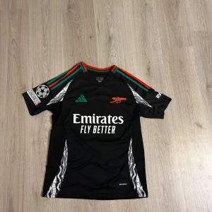 Arsenal Martinelli Champions League tröja - Snygg svart Arsenal fotbollströja från Adidas med Martinelli 11 på ryggen. Tröjan har gröna och röda detaljer på axlarna, Champions League-märke på ärmen och Emirates Fly Better-tryck på bröstet. Tillverkad i lätt och ventilerande polyester.