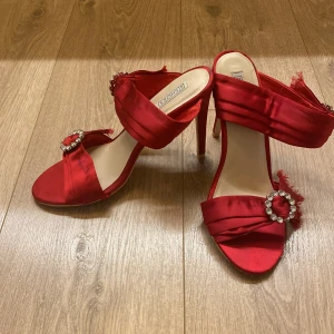 Röda pumps med strassdetalj - Snygga röda pumps med öppen tå och hög klack från Nely Shoes. Skorna är tillverkade i glansig satin och har breda band över foten med en dekorativ strassring framtill. Perfekt för dig som vill sticka ut med färg och bling.