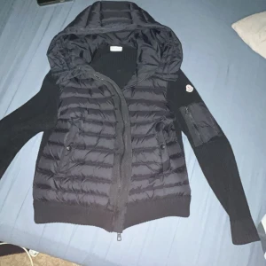 Moncler cardigan  - Svart pufferjacka från Moncler med stickade ärmar och huva. Jackan har quiltad front, två fickor med knappar och en extra ficka med dragkedja på ärmen. Snyggt Moncler-märke på armen och ribbad stickning på ärmar och sidor. Perfekt för dig som gillar streetstyle.