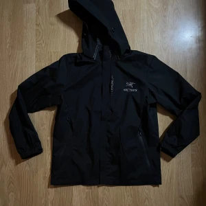 Svart vindjacka från Arc'teryx  - Arc'teryx svart vindjacka med huva och dragkedja framtill. Jackan har två sidofickor med dragkedja och logga på bröstet. Materialet är slitstark syntet som står emot vind och väta, perfekt för utomhusaktiviteter. Snygg och funktionell design med elastiska muddar. Storlek M 