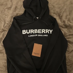 Svart Burberry hoodie med logga - Svart hoodie från Burberry med stor vit logga och texten 'London England' på bröstet. Klassisk huva med snörning och känguruficka framtill. Mjuk och skön, perfekt för en chill look. Säljes i storlek S.