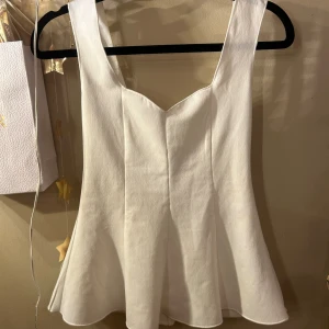 Vit peplumtop från Gina Tricot XS - Supersnygg vit peplumtop från Gina Tricot i storlek XS. Toppen har breda axelband, v-ringad hals och en utsvängd nederdel som ger en feminin siluett. Perfekt att styla med jeans eller kjol för en trendig look.