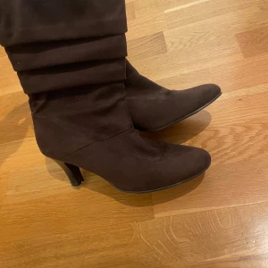 Bruna mockastövletter med klack - Snygga bruna stövletter i mjuk mocka med spetsig tå och medelklack. Skorna har ett veckat skaft som ger en cool och trendig look. Perfekt för dig som gillar att sticka ut med stilrena boots i höst och vinter.