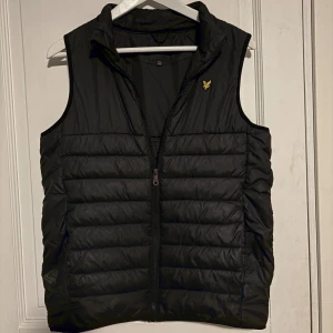 Svart dunväst från Lyle & Scott Junior - Svart dunväst från Lyle & Scott Junior med gul broderad logga på bröstet. Västen har quiltad design, hög krage och dragkedja framtill. Perfekt för lager-på-lager och snygg till både hoodie och t-shirt.