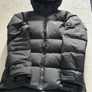 Svart Sail Antarctica dunjacka - Säljer en svart dunjacka från Sail Antarctica Expedition med huva och dragkedja framtill. Jackan har en quiltad design, patch på ärmen och justerbara muddar med kardborre. Perfekt för kalla dagar och riktigt snygg streetstil. Bästa och varmaste vinterjackan jag har haft, jätte bra skick.