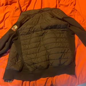 Svart quiltad jacka Parajumpers S - Svart quiltad jacka från Parajumpers med broderad patch på ärmen och dragkedjor vid fickorna. Jackan har en sportig look med ribbade muddar och är tillverkad i fleece och syntetmaterial. Perfekt för dig som gillar streetwear och vill ha något varmt och stilrent.