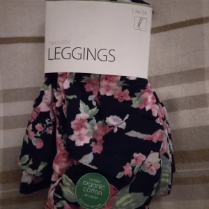 Hely nya, oanvända svarta blommiga leggings från Lindex, Small - Nya oanvända leggings från Lindex i storlek 36/38, tillverkade av ekologisk bomull. De har svart basfärg med blommigt mönster i rosa och grönt. Är medvetet rynkade nedtill. Felfria. Nypris 199 kr.