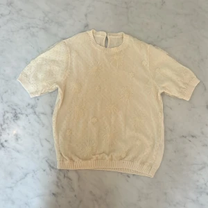 Beige blommig stickad topp från Zara - Supersöt beige stickad topp från Zara med korta ärmar och broderade blommor över hela plagget. Rund halsringning med knapp i nacken och ribbade muddar. 