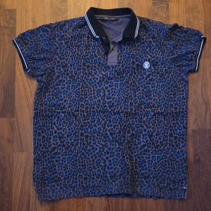Roberto Cavalli - Leopardmönstrad pikétröja - size L - Snygg pikétröja från Roberto Cavalli i svart och blått leopardmönster. Tröjan har klassisk krage med vita ränder, korta ärmar och broderad logga på bröstet. Tillverkad i 100% bomull för en skön känsla. Perfekt för dig som vill sticka ut med stil.