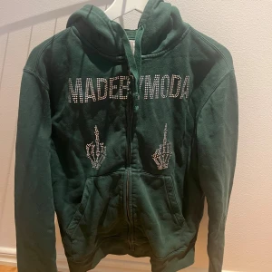 Mörkgrön hoodie MADEBYMODA - Mörkgrön hoodie från MADEBYMODA med dragkedja och huva. Framsidan har stora strasstenar som bildar texten MADEBYMODA och två skelett-händer i strass. Mjuk bomullskänsla och långa ärmar. Perfekt för dig som gillar statement-plagg.