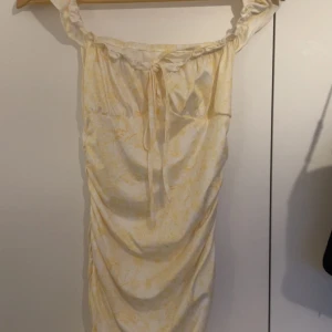 Gulvit offshoulder klänning med snörning - Söt offshoulder klänning i gulvitt med subtilt blommönster. Klänningen har snörning framtill och är figurnära med rynkade detaljer längs sidorna. Perfekt för sommarens varma dagar och har en fräsch, ljus känsla.
