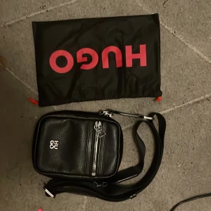 Svart axelväska från HUGO i skinn - Snygg svart axelväska från HUGO i präglat skinn med silverfärgade detaljer och dragkedja framtill. Väskan har justerbar rem och logga både på framsidan och på dragkedjan. Perfekt för att bära det viktigaste och ger en clean look.