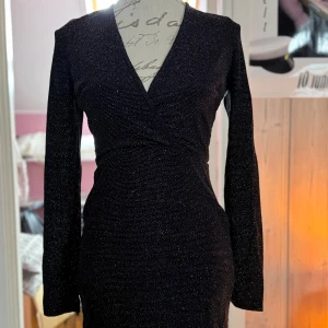 Glittrig svart klänning från NLY One - Supersnygg svart glittrig klänning från NLY One med lång ärm och v-ringad omlottfront. Tight passform som framhäver figuren och skimrande material som fångar ljuset. Perfekt för dig som vill sticka ut med stil.