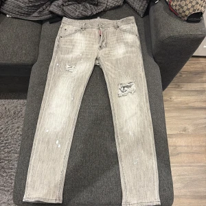 Grå Dsquared2 jeans med slitningar - Säljer ett par grå jeans från Dsquared2 med coola slitningar och färgstänk på benen. Jeansen har rak passform, klassisk femficksdesign och tydlig Dsquared2-logga vid fickan. Perfekta för dig som gillar en edgy streetstil.