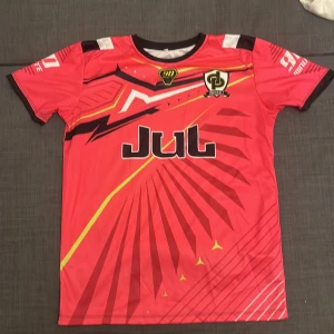 Röd fotbollströja JUL x Yamaha - Snygg röd fotbollströja från 90 Minute med JUL-logga och Yamaha-tryck på ryggen. Tröjan har coola grafiska detaljer i svart, vitt och gult, samt klubbmärke 'Don De Platine' på bröstet. Kortärmad modell i lätt polyester, perfekt för match eller träning.