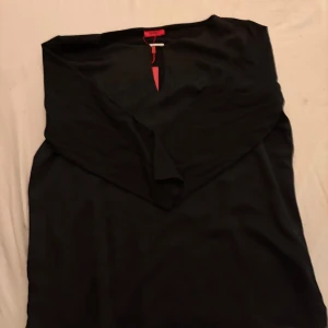 Svart blus från Hugo Boss - En stilren svart blus från Hugo Boss med v-ringad hals och lång ärm. Klänningen har en enkel, elegant design och är tillverkad i ett mjukt tyg som faller snyggt. Perfekt för dig som gillar klassisk och tidlös stil.