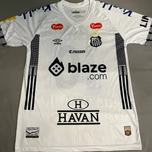 Vit Santos FC Neymar Jr. matchtröja - Säljer en officiell vit Santos FC fotbollströja från Umbro med Neymar Jr. och nummer 10 på ryggen. Tröjan har korta ärmar, flera sponsorer tryckta fram och bak, samt klubbmärket på bröstet. Materialet är lätt och andas, perfekt för träning eller match.