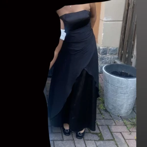 Svart långklänning Juju & Christine - Elegant svart långklänning från Juju & Christine med omlott-detalj framtill och snörning i ryggen. Klänningen är axelbandslös och har en stilren, figurnära passform. Materialet är glansigt och känns lyxigt, perfekt för dig som vill sticka ut med en klassisk look. Endast använd en gång.