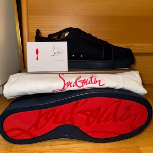 Christian Louboutin Mörkblå sneakers - Snygga svarta sneakers från Christian Louboutin i mocka med klassisk röd sula och diskreta nitar på sidan. Skorna har rund tå, platt sula och snörning. Kommer med originalkartong, dustbag och äkthetskort. Perfekta för dig som vill sticka ut med exklusiv stil.