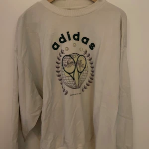 Vit Adidas sweatshirt med tennistryck kommer i sett med short - Vit sweatshirt från Adidas med grönt tryck framtill som visar två tennisrack och texten 'Tennis Academy'. Tröjan har rund halsringning, långa ärmar och ribbade muddar. Adidas-logga i grönt på ryggen. Perfekt för en sportig och avslappnad look. Även shorts som är i samma modell