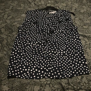 Svart prickig bluse från Flash Woman - Snygg svart bluse från Flash Woman med vita prickar och volangdetaljer framtill. Blusen är ärmlös och har en lätt A-linjeformad siluett som ger en skön och luftig känsla. Perfekt för dig som gillar lekfulla mönster och vill sticka ut.