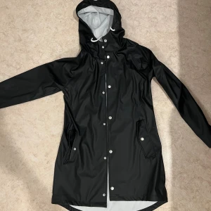 Svart regnjacka från 157 - Svart regnjacka från 157 i storlek XS. Jackan har huva med vit snörning, långa ärmar och knäppning med silvriga tryckknappar framtill. Jag köpte dom för 300kr och prislappen är kvar. Den är för liten för mig och kunde ej lämna tillbaka eftersom det hade gått ut på öppet köp, därför säljer jag den! 