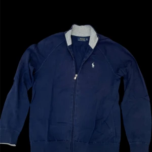 Marinblå zip-kofta från Polo Ralph Lauren - Snygg marinblå kofta från Polo Ralph Lauren med dragkedja hela vägen och grå ribbad krage. Klassisk broderad logga på bröstet och mjukt bomullsmaterial. Perfekt för dig som gillar stilrena och sportiga plagg. Pris kan diskuteras 