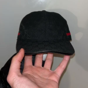 Svart Gucci keps med GG-mönster - Svart keps från Gucci med diskret GG-monogram över hela ytan och klassiska gröna och röda ränder på sidorna. Tillverkad i Italien och gjord av bomull. Snygg böjd skärm och justerbar passform. Perfekt för dig som vill ha en lyxig streetwear-look.