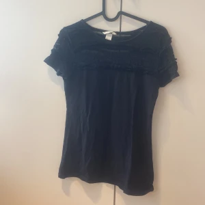 Svart topp med spets från H&M - Svart topp från H&M i storlek S med korta ärmar. Överdelen har snygga spetsdetaljer och diskreta volanger som ger en cool och feminin touch. Mjuk och skön bomullskänsla i materialet, perfekt till jeans eller kjol.