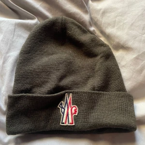 Mörkgrön Moncler mössa med logga - Snygg mörkgrön mössa från Moncler Grenoble med broderad logga i rött, vitt och blått framtill. Mössan är stickad och har en uppvikt kant för extra stilpoäng. Perfekt för dig som vill ha en clean look med exklusiv känsla.