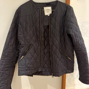 Svart quiltad jacka från Boomerang - Quiltad svart jacka från Boomerang med snygga guldfärgade dragkedjor på fickor och ärmslut. Jackan har rund hals, lång ärm och ett stilrent rutmönster. Perfekt för dig som gillar klassisk men modern look.