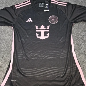 Inter Miami CF svart fotbollströja Adidas - Säljer en officiell Inter Miami CF fotbollströja från Adidas i svart med rosa detaljer och klubbmärke på bröstet. Tröjan har korta ärmar, rosa ränder på axlarna och är tillverkad i lätt, ventilerande AEROREADY-material. Perfekt för dig som älskar fotboll och snygg design.