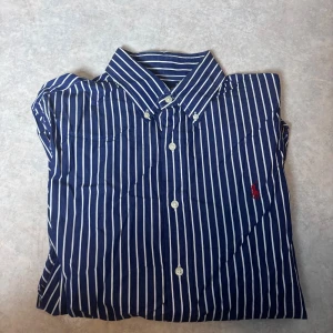 Blåvit randig skjorta Ralph Lauren M - Snygg blå skjorta med vita vertikala ränder från Ralph Lauren, slim fit-modell. Klassisk krage, långa ärmar och knappar framtill. Ikonisk röd Polo-logga broderad på bröstet. Perfekt för dig som gillar stilrena och tidlösa plagg.