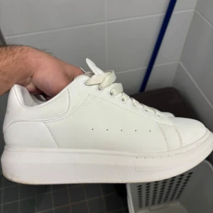 Vita Alexander McQueen sneakers - Säljer ett par stilrena vita sneakers från Alexander McQueen med chunky plattformssula och rund tå. Skorna har snörning och är tillverkade i skinn med diskreta gulddetaljer på insidan. Perfekta för dig som gillar minimalistisk och lyxig streetstil.