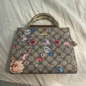 Blommig handväska från Gallantry - Snygg handväska från Gallantry med blommigt mönster i rosa, blått och rött på grå botten. Väskan har guldfärgade handtag och detaljer samt flera fack inuti med dragkedja. Materialet är syntet med inslag av brunt konstläder på insidan.