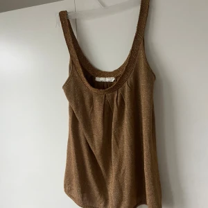 Glittrig brun/guld linnetopp från Mango - Snygg brun linnetopp från Mango med glittrigt tyg och breda axelband. Toppen har en djup rundad ringning och ett lätt fall i materialet. Perfekt för dig som vill sticka ut med en touch av glitter.