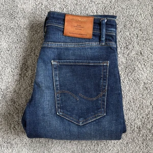 Jack & Jones blå jeans Intelligence - Snygga blå jeans från Jack & Jones Intelligence med klassisk femficksdesign och kontrastsömmar. Jeansen har en mörk tvätt med lätt slitning och bruna detaljer på läderpatchen. Passar dig som gillar stilrena och tidlösa jeans med bekväm passform.