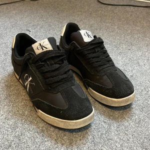 Svarta sneakers Calvin Klein Jeans - Svarta sneakers från Calvin Klein Jeans med vit logga på sidan och vit detalj på hälen. Skorna har snörning, platt sula och rund tå. Materialmix av syntet och mocka ger en cool kontrast. Perfekt för dig som gillar stilrena och klassiska sneakers. Nypris 1400 pris kan diskuteras.