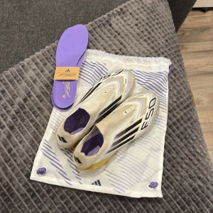 Adidas F50 fotbollsskor vit/guld - Säljer ett par Adidas F50 fotbollsskor i vitt med svarta och guldiga detaljer. Skorna har en strömlinjeformad design, platt sula med dobbar i guld och svart F50-tryck på ovansidan. Innersulan är lila och medföljer en matchande vit och lila påse. Perfekt för dig som vill sticka ut på planen. Andvänd 2 gånger, storlek 43 