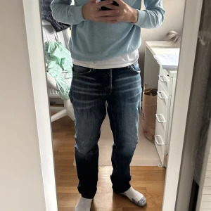 Blå jeansbyxor med straight fit - Säljer ett par blå jeansbyxor med snygga slitningar och tvättade detaljer. Modellen har rak passform och klassisk femficksdesign. Jeansen har normal midja och är tillverkade i robust denim. Perfekt för dig som gillar en avslappnad och stilren look.