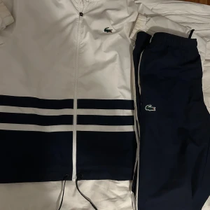 Lacoste tracksuit/träningsdress - Säljer en snygg tracksuit från Lacoste i vitt och marinblått. Jackan har dragkedja,  Byxorna är marinblå med vit sidopanel och klassisk Lacoste-logga. Perfekt för sportiga dagar eller chill.  tracksuiten i färgen mörkblå och vit som sitter perfekt på än. Den kan användas året runt och går för 2800 kr i affär, i storlek S men passar även någon som precis kommit in i storlek M pris kan diskuteras så tveka inte på att höra av er