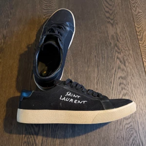 Svarta Saint Laurent sneakers - Snygga svarta sneakers från Saint Laurent med vit handskriven logga på sidan. Skorna har svart canvas och detaljer i skinn, samt en beige platt gummisula. Klassisk rund tå och snörning framtill. Perfekta för dig som gillar stilrena och exklusiva sneakers.