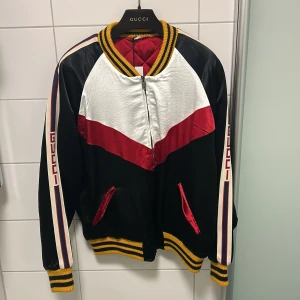 Gucci bomberjacka i svart, vitt och rött storlek 50 - Snygg bomberjacka från Gucci med svart, vitt och rött i färgblock. Jackan har gula och svarta ribbade muddar, dragkedja framtill och röda detaljer. Ärmarna har Gucci-logga och ränder i vitt, rött och blått. Materialet är glansigt och jackan är quiltad på insidan. Helt ny 