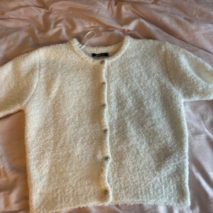 Vit kortärmad kofta från Gina tricot  - Supermjuk och fluffig vit kofta från Gina tricotmed korta ärmar och rund hals. Koftan har knappar framtill och är gjord i ett mysigt, stickat material som känns extra cozy. Perfekt för lager-på-lager och chill vibes. Finns en liten fläck längst ner på framsidan 