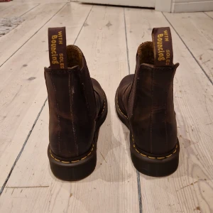 Bruna Chelsea boots Dr. Martens - Säljer ett par bruna Chelsea boots från Dr. Martens i läder med klassisk gul söm runt sulan och elastiska paneler på sidorna. Dragflikar med gul text baktill. Perfekta för dig som gillar tidlös och robust stil. De är inte använda någon gång