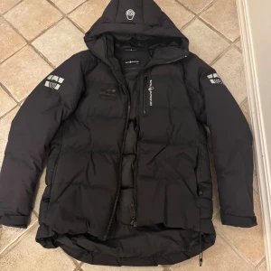 Svart dunjacka från Sail Racing - Säljer en svart dunjacka från Sail Racing med huva och feta loggor på ärmarna och bröstet. Jackan är vadderad med 500 FP dun från Allied Feather + Down och har dragkedja framtill samt flera fickor. Perfekt för kalla dagar när du vill hålla stilen. Bara en av dragkedjorna är bytta. 