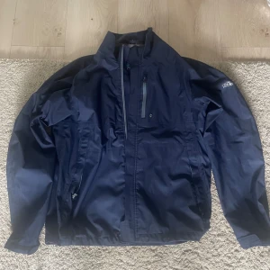 Mörkblå vindjacka från Oscar Jacobson - Säljer en mörkblå vindjacka från Oscar Jacobson med hög krage och dragkedja framtill. Jackan har en bröstficka med dragkedja och två sidofickor. Materialet är syntet och jackan är perfekt för blåsiga dagar. Enkel och stilren design med logga på ärmen.