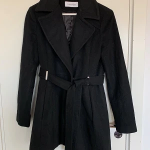 Svart kappa från Calvin Klein med bälte - Säljer en svart kappa från Calvin Klein med klassisk krage och bälte i midjan. Jackan har långa ärmar, knappar på insidan och är fodrad för extra komfort. Perfekt för dig som vill ha en stilren och tidlös look.