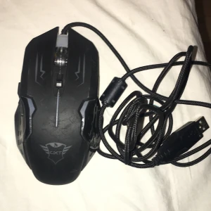Trust GXT Gaming Mouse - Ergonomisk Trust GXT gamingmus med USB-anslutning och flätad kabel för extra hållbarhet. Optisk sensor för precis styrning, flera knappar och gamingdesign. Mindre bruksspår syns men i gott skick. Perfekt för gaming eller arbete.